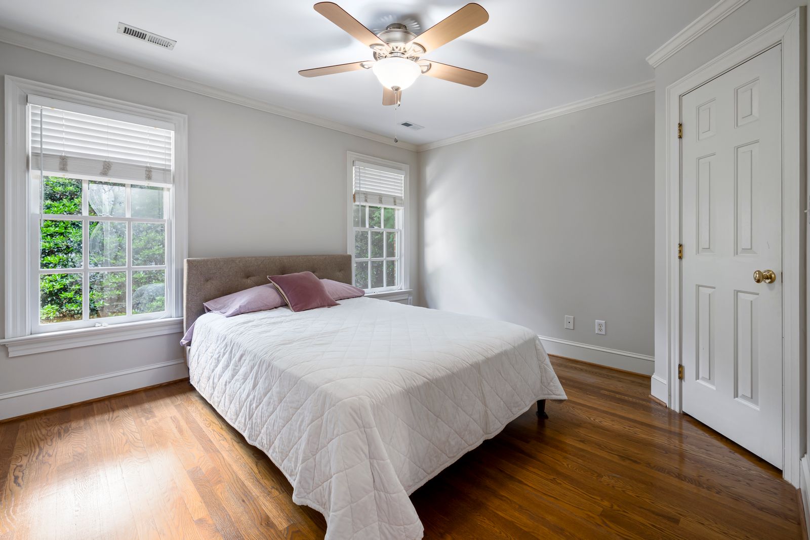 A bright, simply-furnished bedroom with a white quilted bed, ceiling fan, wood floors, and windows looking onto greenery